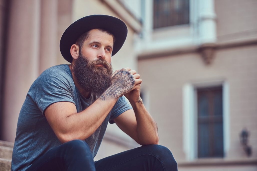 Handsome hipster male with a stylish beard with a tattoo on his arms dressed in casual clothes and hat sits on steps.